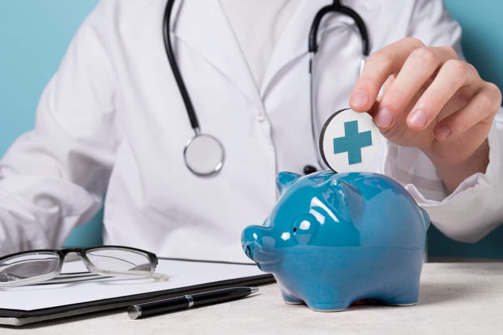 a doctor in medical white suit putting a coin with croos into blue piggy bank